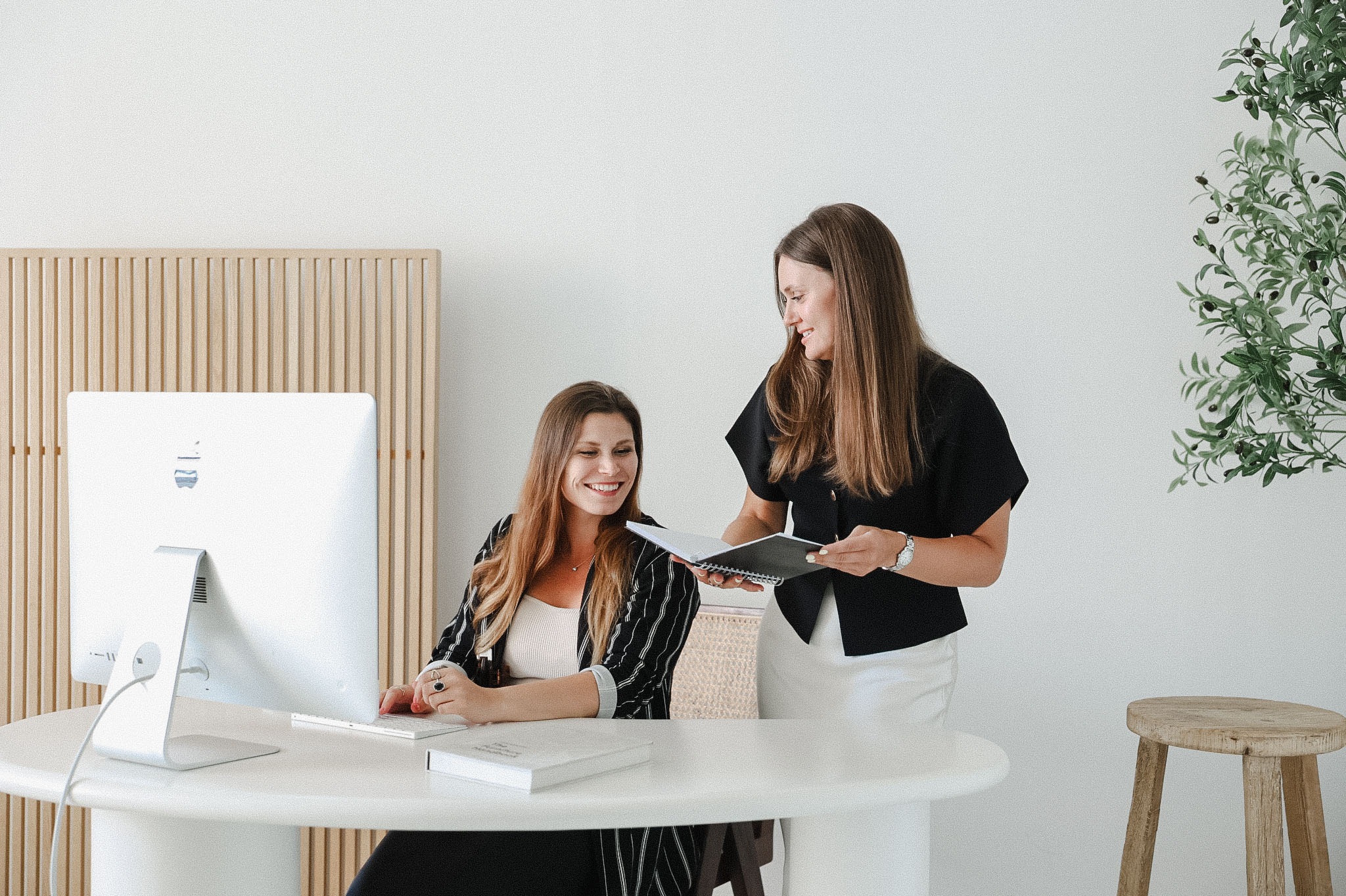 Two women in business attire work together at a desk with a computer; one is seated and smiling, while the other stands holding a clipboard and papers—showcasing effective marketing on a budget.