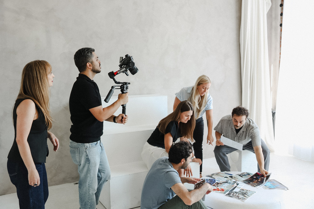 Six people collaborate around a small table with magazines, brainstorming budget marketing ideas, while one person films the group using a stabilizer-mounted camera in a bright, minimalistic room.