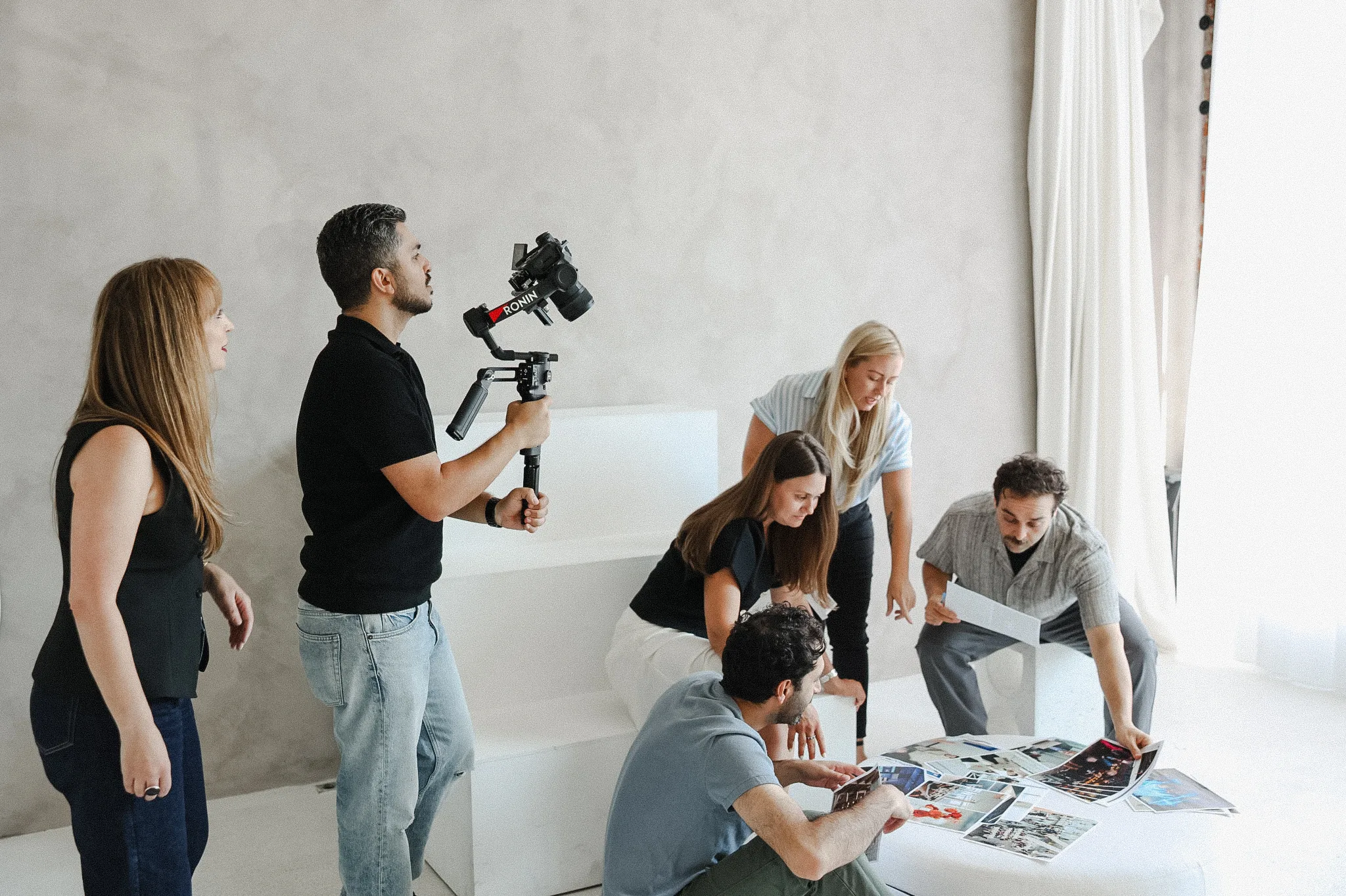 Six people collaborate around a small table with magazines, brainstorming budget marketing ideas, while one person films the group using a stabilizer-mounted camera in a bright, minimalistic room.