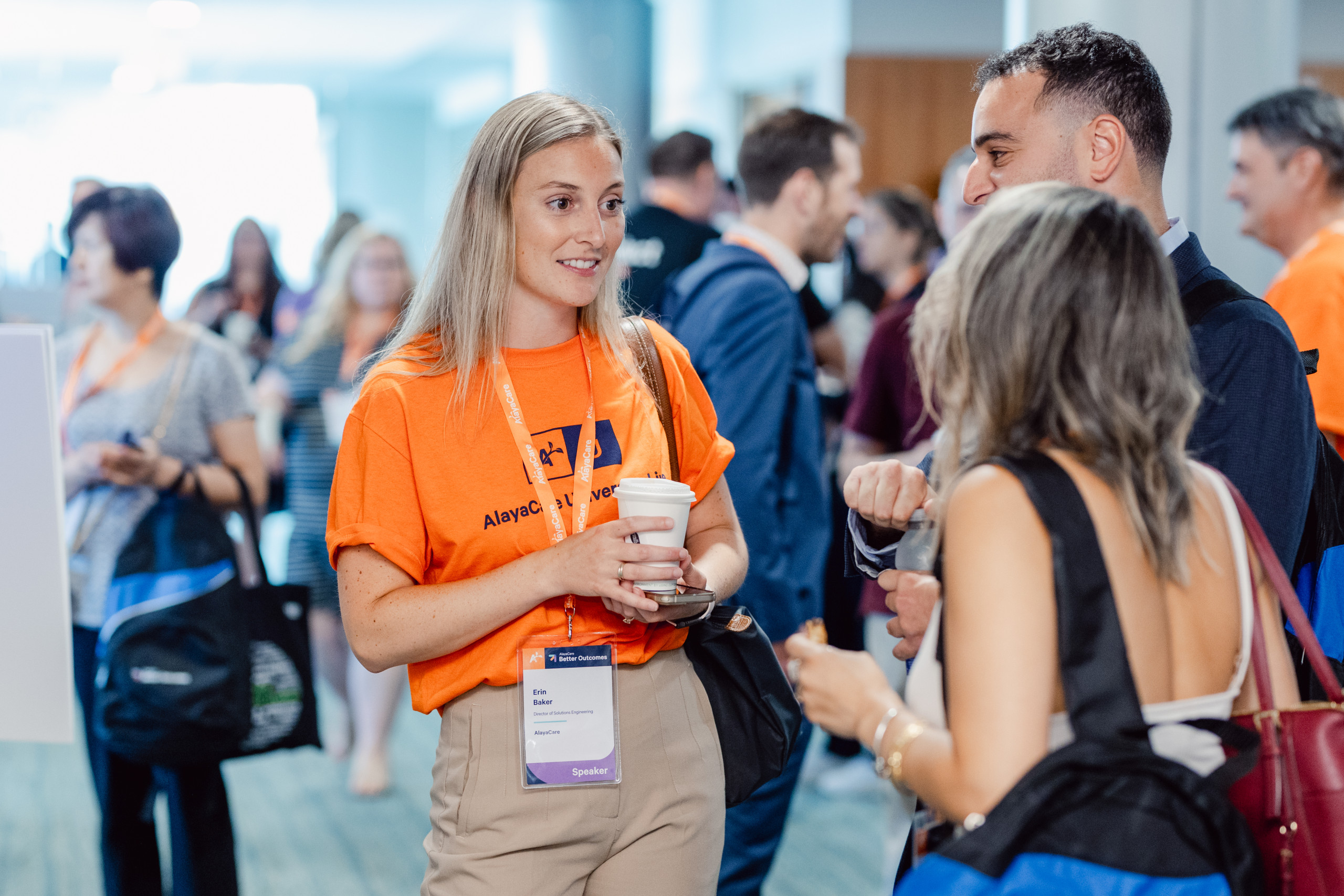 A woman in an orange shirt talks with two people at a conference; she holds a coffee cup and wears a name badge, with other attendees visible in the background.