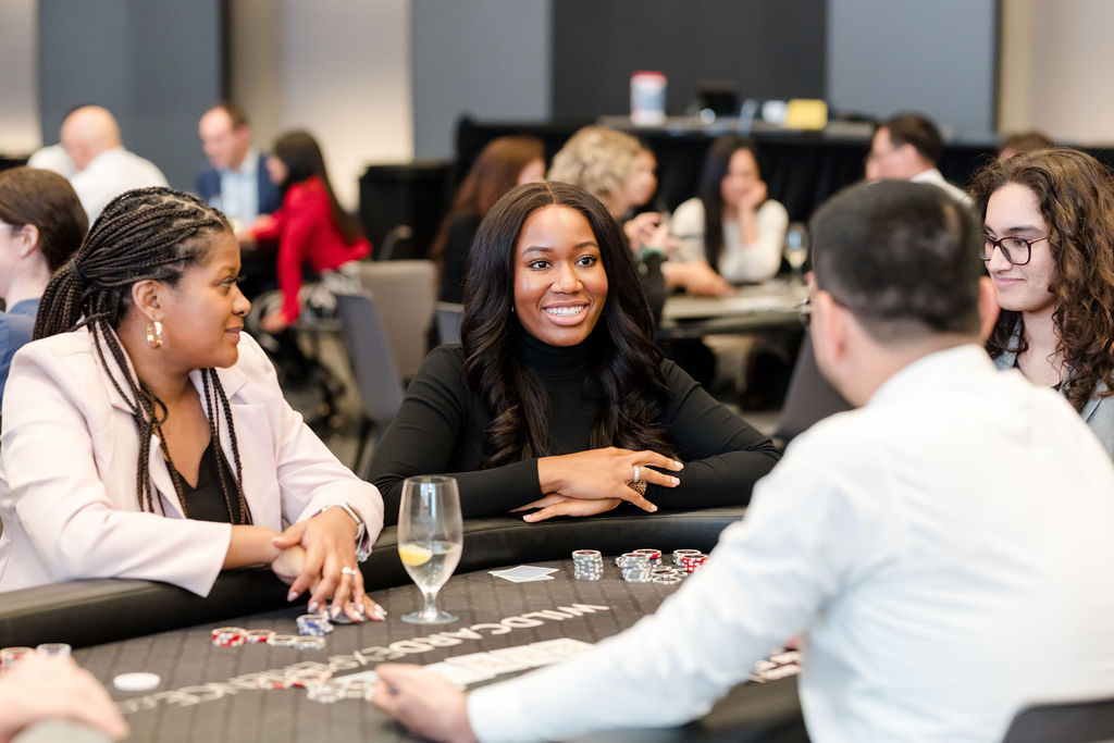 People sit around a blackjack table engaged in conversation, with poker chips and a glass of water on the table. The setting appears to be a social or corporate event.
