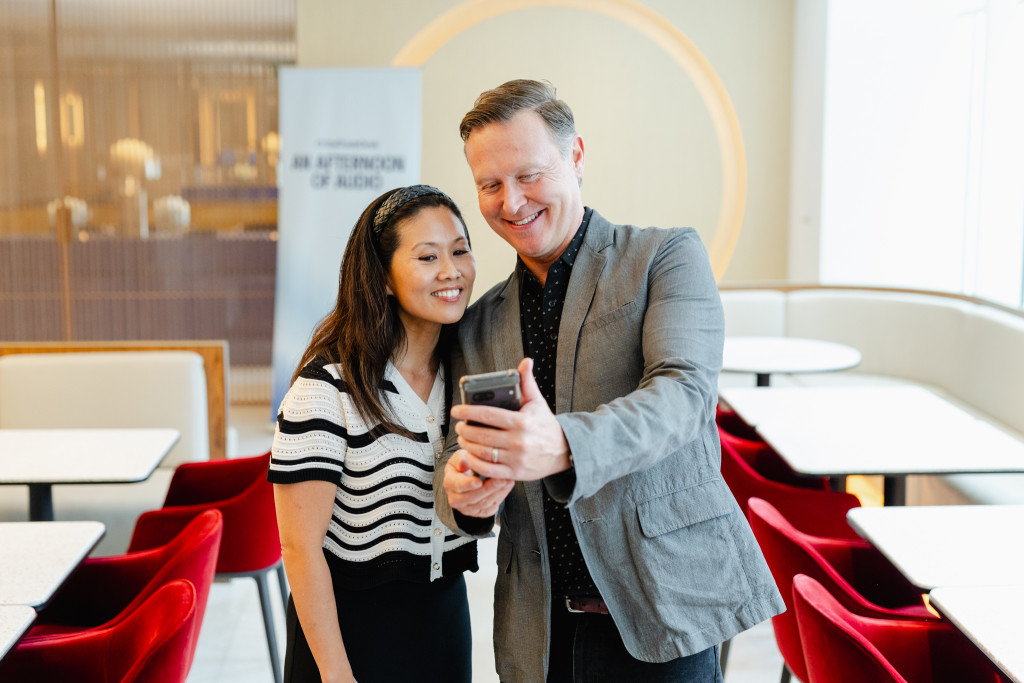 A man and a woman stand close together, smiling and looking at a smartphone in a modern, well-lit cafe with red chairs and white tables, perhaps discussing high-impact content or marketing on a budget.