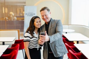 A man and a woman stand close together, smiling and looking at a smartphone in a modern, well-lit cafe with red chairs and white tables, perhaps discussing high-impact content or marketing on a budget.