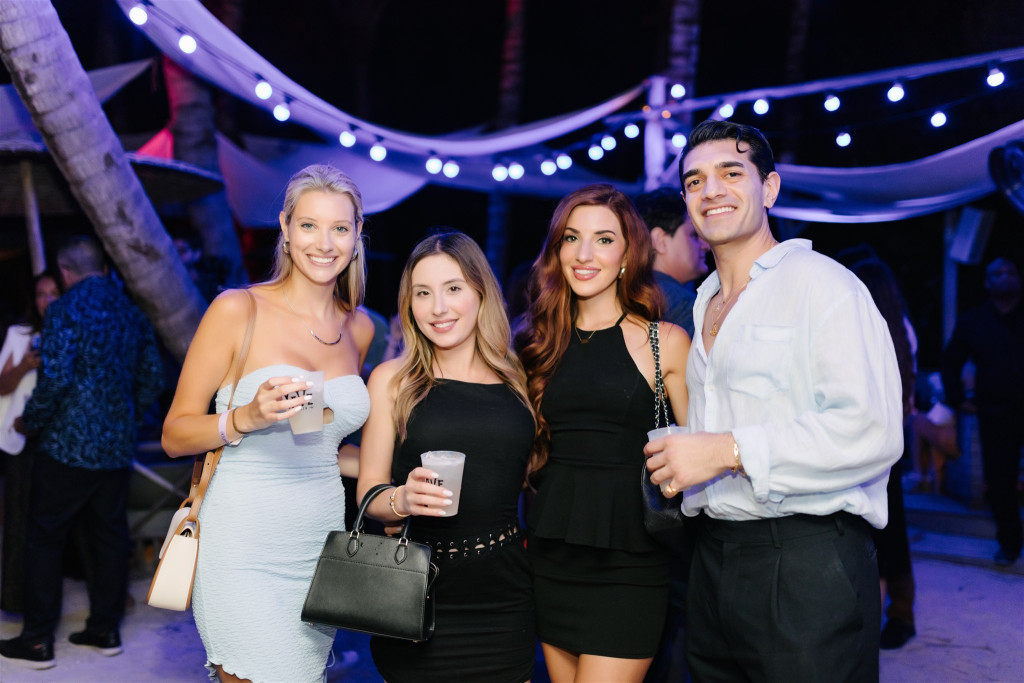 Four people stand together at an outdoor nighttime event, holding drinks and smiling, with string lights and decorations visible in the background.
