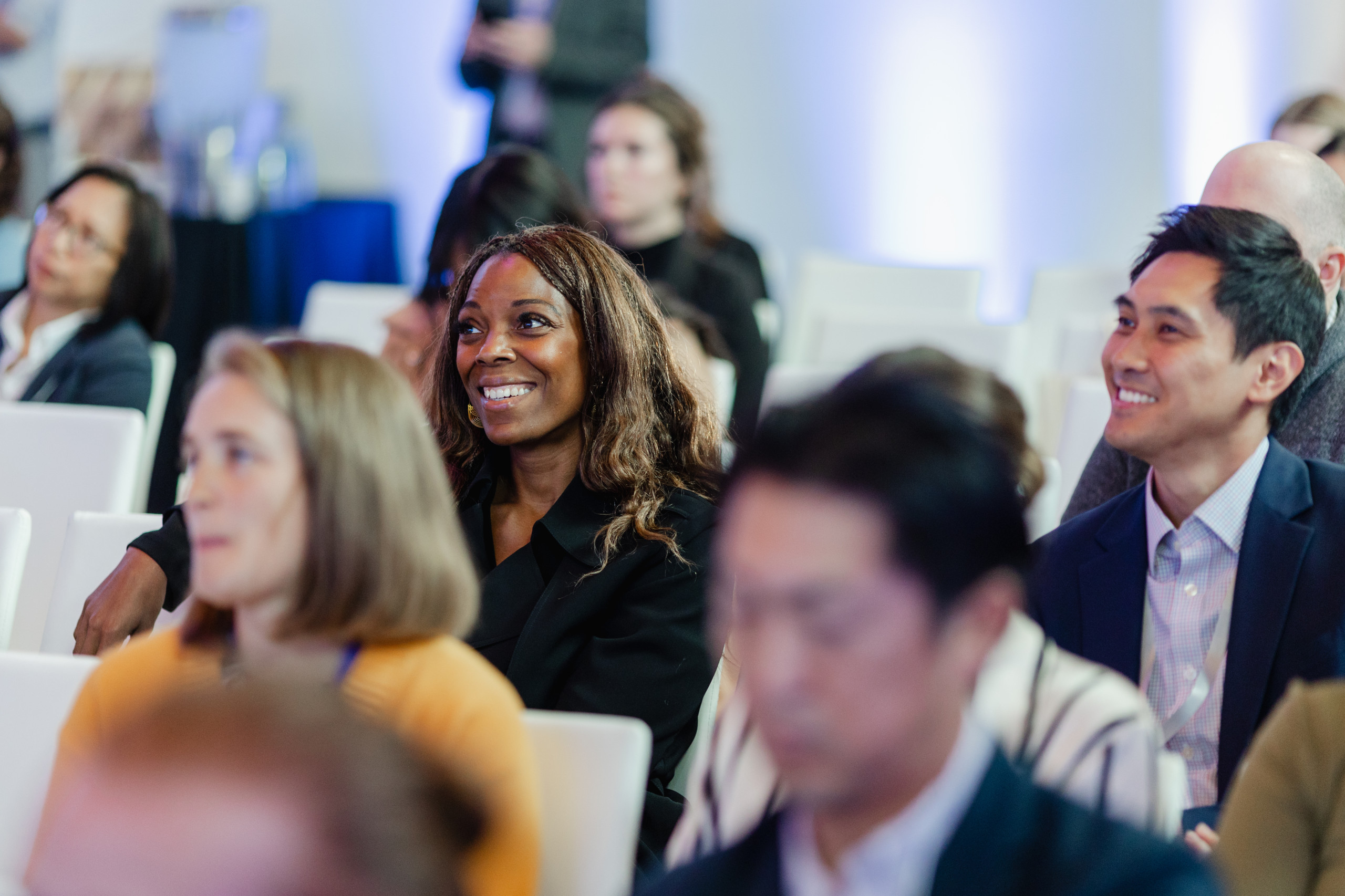 A group of people sit in an audience, attentively watching an event. Several individuals are smiling and dressed in business attire.