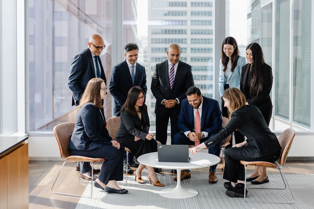 A group of business professionals in formal attire gathers around a laptop in a modern office with large windows and city views, collaborating on high-impact content strategies to navigate a downturn.
