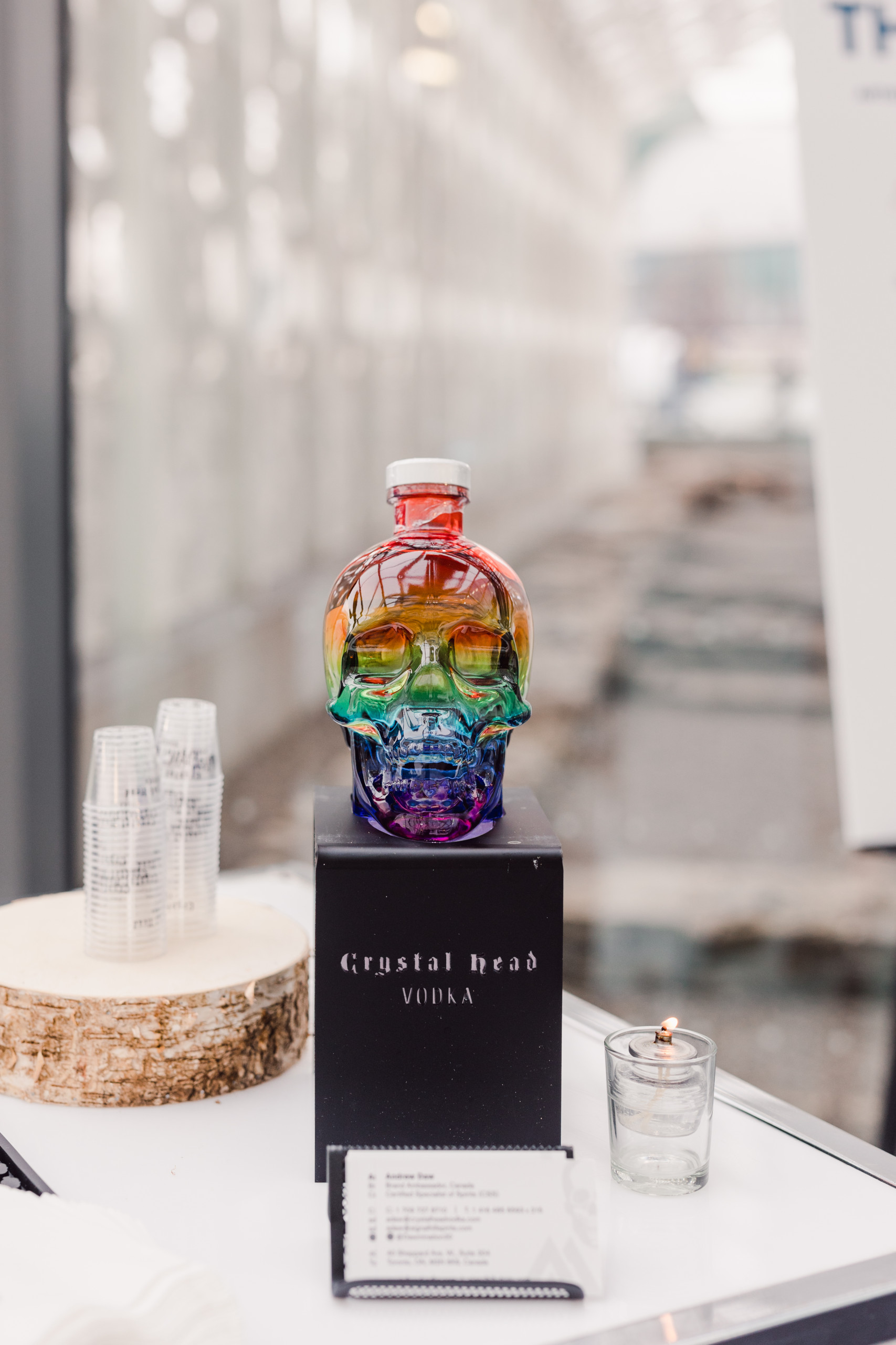 A rainbow-colored Crystal Head Vodka skull bottle is displayed on a black stand next to plastic cups, a candle, and a drink menu on a white surface.