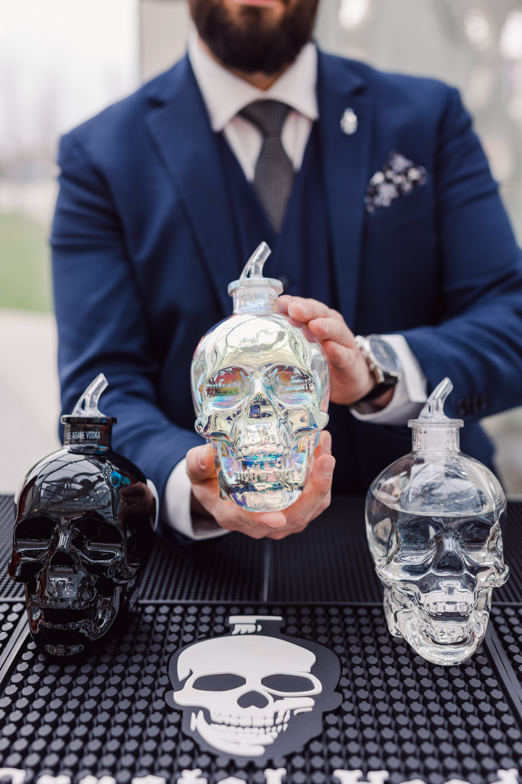 A man in a blue suit holds a clear, iridescent skull-shaped bottle between a black and a clear skull bottle on a bar mat with a skull graphic.