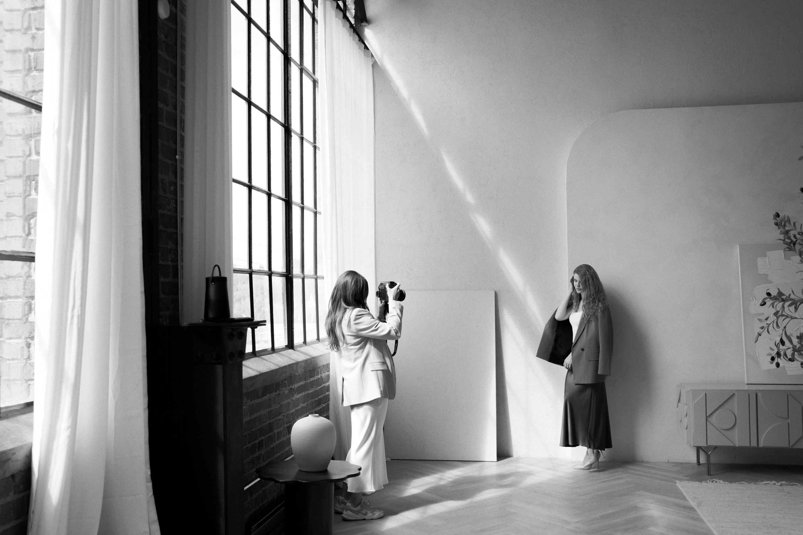 A woman takes a photo of another woman standing against a wall during a content shoot in a well-lit, minimalist room with large windows and modern decor.