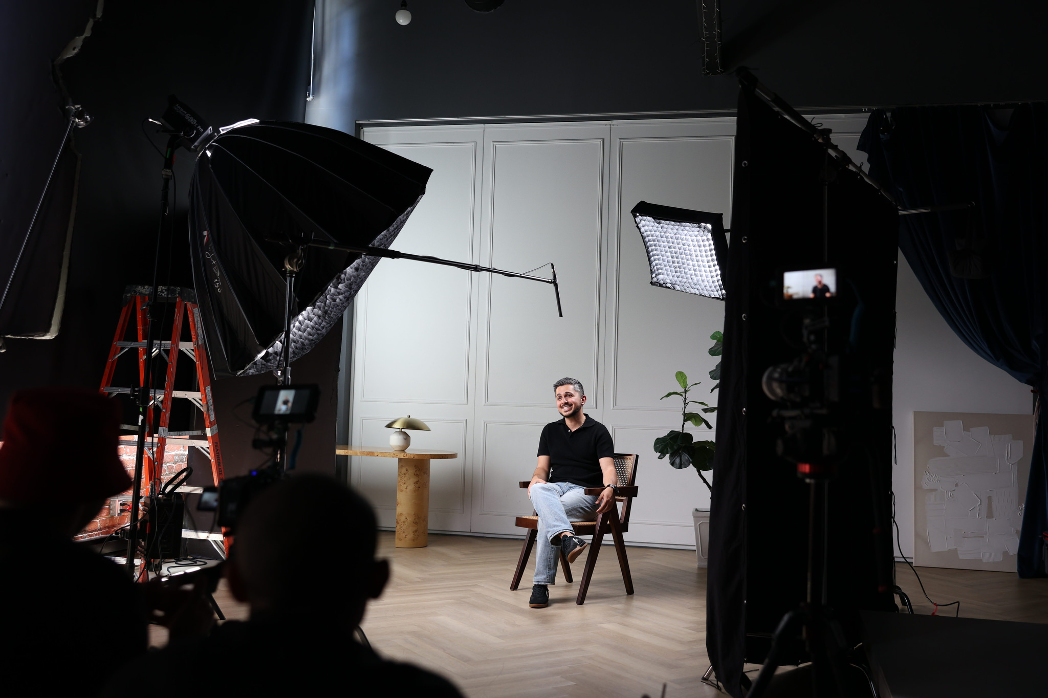 A man sits on a chair in a photography studio during a content shoot, surrounded by cameras, lights, and equipment, with a white wall and furniture in the background.
