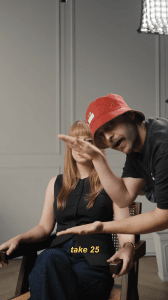 A man in a red bucket hat gestures while a woman with long red hair sits beside him. Yellow text at the bottom reads, "take 25.